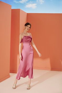 a woman in a pink dress standing against an orange wall with her hand on her hip