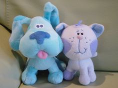 two blue and white stuffed animals sitting next to each other on a couch, one with its tongue out