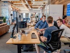 three men sitting at a desk in an office working on their laptops and computers