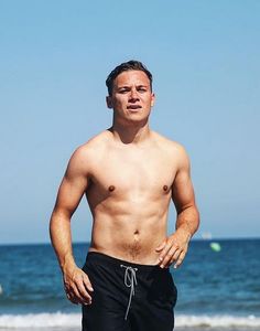 a shirtless man standing on the beach with his hands in his pockets and looking at the camera