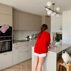 a woman in red shirt standing at counter next to oven and stove top with cat sitting on stool