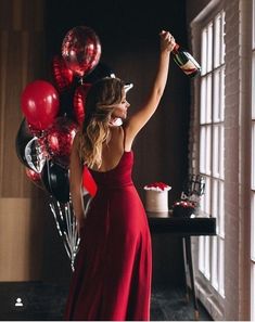 a woman in a red dress is holding a bottle and some black and red balloons