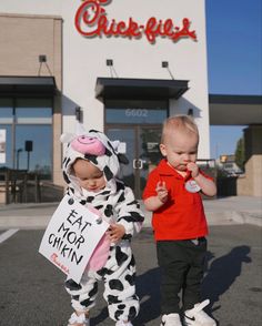 two toddlers dressed as cows standing in front of a chicken restaurant and holding signs