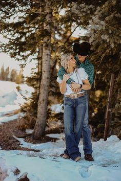 a man and woman standing in the snow near some trees with their arms around each other