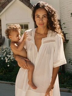 a woman holding a baby in front of a white house with flowers on the side walk