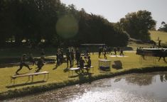 a group of people are running around in the grass next to a river and picnic tables