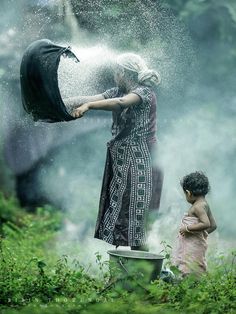 a woman and child standing in the grass with water pouring from an open fire hydrant