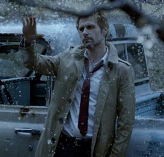 a man in a trench coat and tie standing next to a car on a rainy day