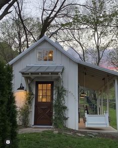 a small white house with a porch and swing set in front of the door, surrounded by trees