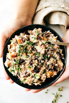 a person holding a black bowl filled with oatmeal and vegetables on top of a white table