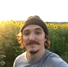 a man with a moustache standing in front of a field of sunflowers