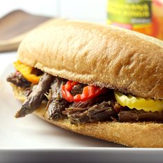 a roast beef sandwich with peppers and onions on a white plate next to a bottle of mustard