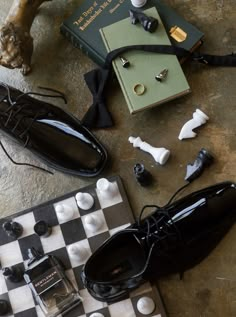 An artistic arrangement of groom accessories including glossy black dress shoes, a black bow tie, cufflinks, a wedding band, and a luxury cologne, all placed on a chessboard with vintage books, symbolizing elegance and sophistication for a luxury wedding. Wedding Accessory Photos, Groom Detail Shots, Groom Idea, Sailboat Elopement, Creative Flatlay, Groom Shoot, Nyc Editorial, Weddings 2024, Groom Prep