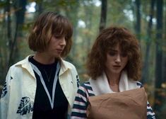 two young women walking in the woods with one holding a paper bag on her shoulder