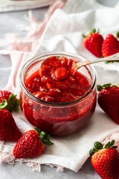 strawberry jam in a glass jar with strawberries around it on a white tablecloth