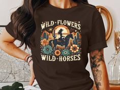 This graphic T-shirt features a unique Wild Flowers Wild Horses design, perfect for anyone who loves vintage western themes and boho styles. Showcasing a retro desert scenery with floral elements and a cowboy rider, it adds a touch of rodeo charm to your wardrobe. Ideal for casual outings or themed events, this shirt is a great addition to any collection.  WELCOME TO RAIN APPAREL US  We are a small business located in Missouri. We started making t-shirts in 2019 and have grown so much since then! We take pride in the quality of our products and are always striving to grow. We hope you can find whatever you are looking for in our shop. Please reach out to our team if you need any help at all! Quality Assurance: We pride ourselves on using high-quality fabrics and safe inks. Our suppliers as Bohemian Graphic Print Rodeo Top, Bohemian Graphic Print Tops For Rodeo, Retro Crew Neck Tops For Western-themed Events, Graphic Print Short Sleeve T-shirt For Country Events, Vintage Letter Print Tops For Western-themed Events, Retro Summer T-shirt For Western-themed Events, Retro T-shirt For Western-themed Summer Events, Graphic Print Crew Neck T-shirt For Country Events, Vintage Short Sleeve Tops For Western-themed Events