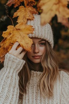 a woman with long hair wearing a white hat and sweater holds up a yellow leaf in front of her face