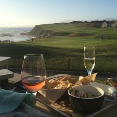 two glasses of wine and some food on a table near the ocean with people in the background