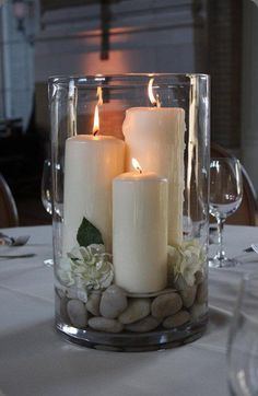 three white candles are sitting in a glass vase with rocks and greenery on the table