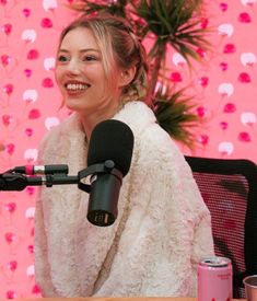 a woman sitting at a table with a microphone in front of her and a pink wall behind her