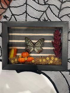 a shadow frame with a butterfly and autumn decorations in it on a black table cloth