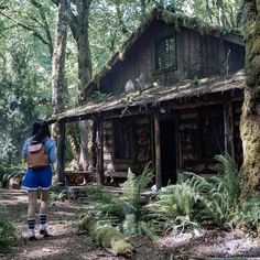 a woman with a backpack is standing in front of a log cabin surrounded by trees and ferns