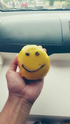 a hand holding a yellow smiley face doughnut in front of a car dash board