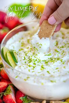 a hand dipping a cracker into a bowl of dip with strawberries and limes in the background