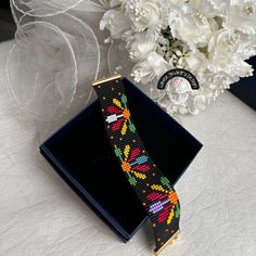 a cross stitch bracelet sitting on top of a black box next to a bouquet of white flowers