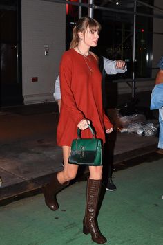 a woman walking down the street carrying a green handbag and wearing knee high boots