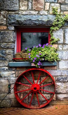 a red wagon with flowers in it sitting on the side of a brick building next to a window