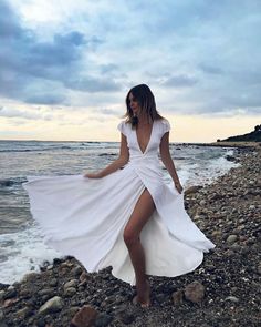 a woman in a white dress is standing on the rocks by the ocean with her legs spread out