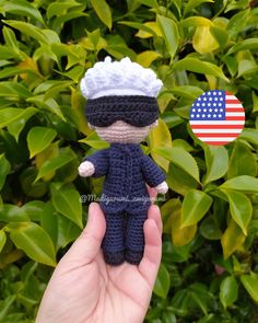 a small crocheted doll with an american flag on it's head is held in front of some bushes