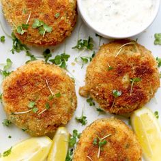 three crab cakes with lemon wedges and parsley on a white plate next to a small bowl of ranch dressing