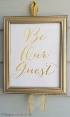 a gold framed sign that says be our guest hanging on the wall with a ribbon