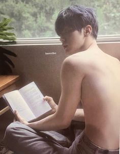 a shirtless young man sitting on a bench reading a book with trees in the background