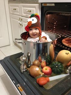 a baby wearing a turkey hat sitting in a pot full of vegetables and apples on the stove