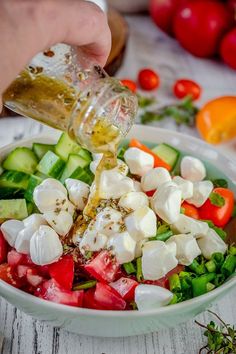 a person pouring dressing into a bowl of salad with tomatoes, cucumbers and other vegetables