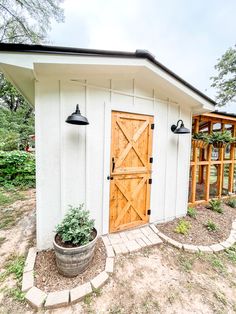 a small white shed with a wooden door and two planters on the front porch