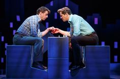 two men sitting on stools facing each other in front of a black background with purple squares