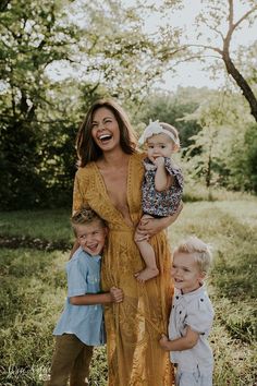 a woman in a yellow dress holding two children and smiling at the camera with trees behind her