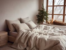 an unmade bed sitting in front of a window next to a small christmas tree