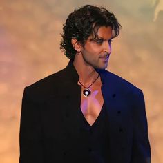 a man in a black suit and necklace looking off to the side with his right hand on his hip