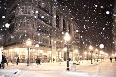 snow falling on the ground in front of an old building at night with people walking around