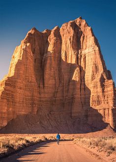 a person walking down a dirt road in front of a large rock formation on the side of a mountain