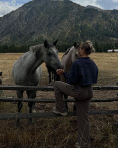 a woman standing next to a fence with two horses in the field behind her and mountains in the background