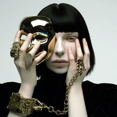 a woman with black hair and gold jewelry holding up a mask to her face while looking at the camera