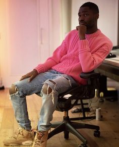 a man in a pink sweater and ripped jeans sits on an office chair