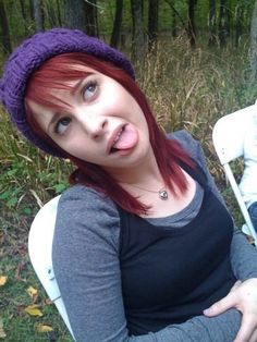 a woman with red hair sitting on a white chair in the woods wearing a purple knitted hat