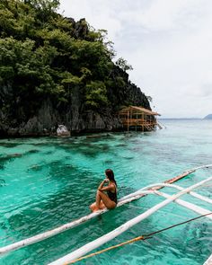 a woman sitting on the bow of a boat in clear blue water next to an island